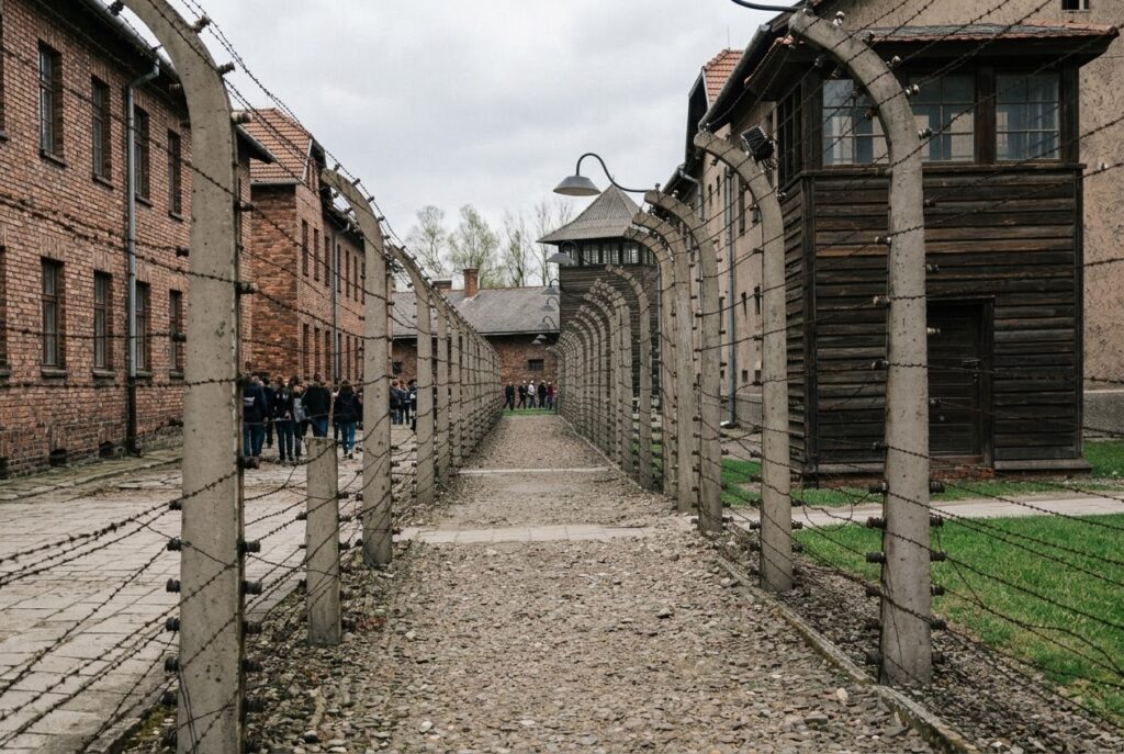 Double barbed-wire fences and brick barracks at the Auschwitz II-Birkenau memorial on an overcast day, with visitors walking the historic gravel paths